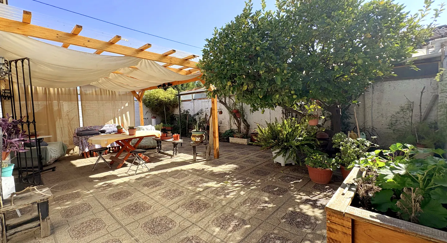 Outdoor patio with a wooden pergola covered by white fabric, a red table, and various plants in pots. The ground is tiled.