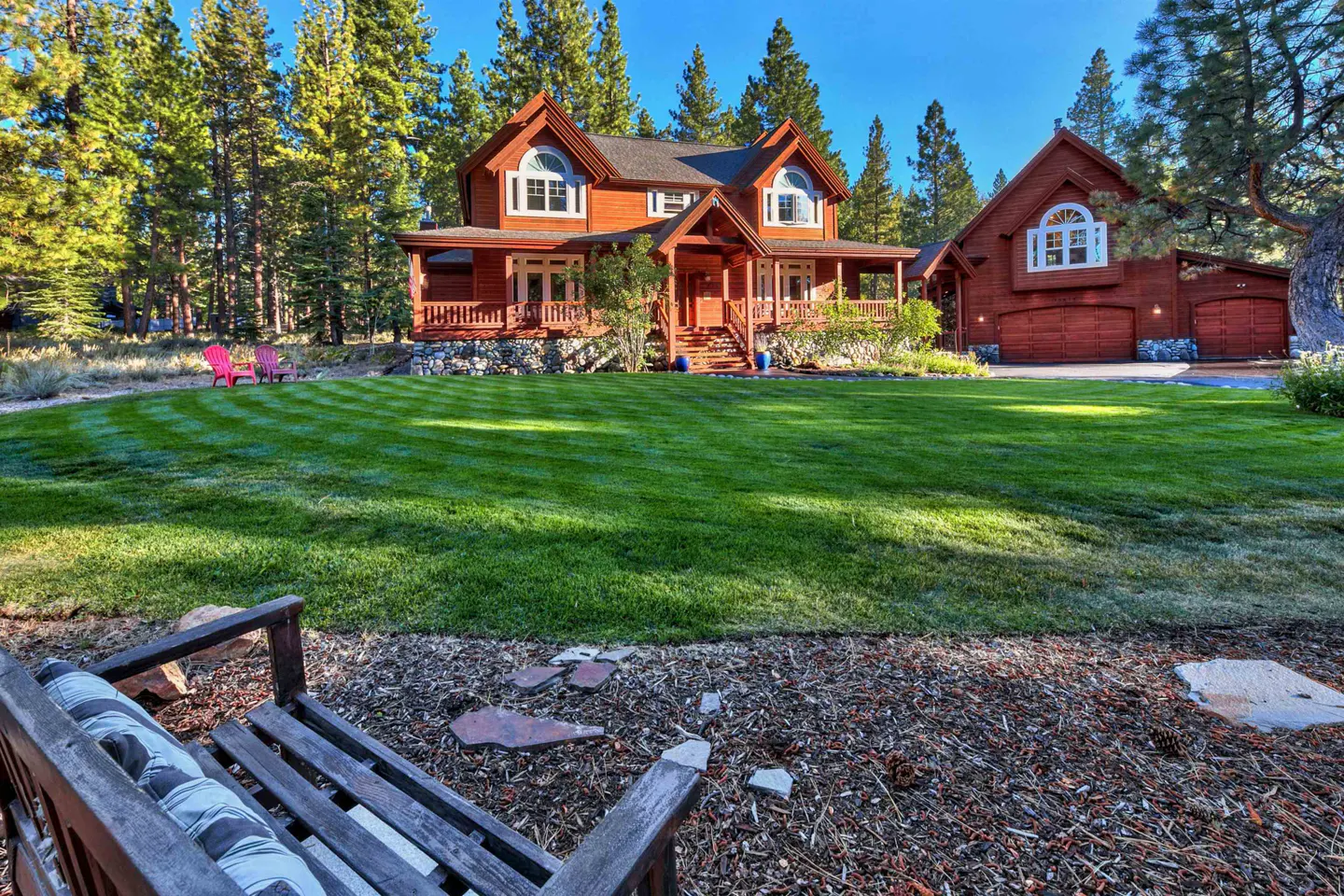 Exterior of a two-story wood house with a green lawn and tall trees. A wooden bench sits in the foreground.