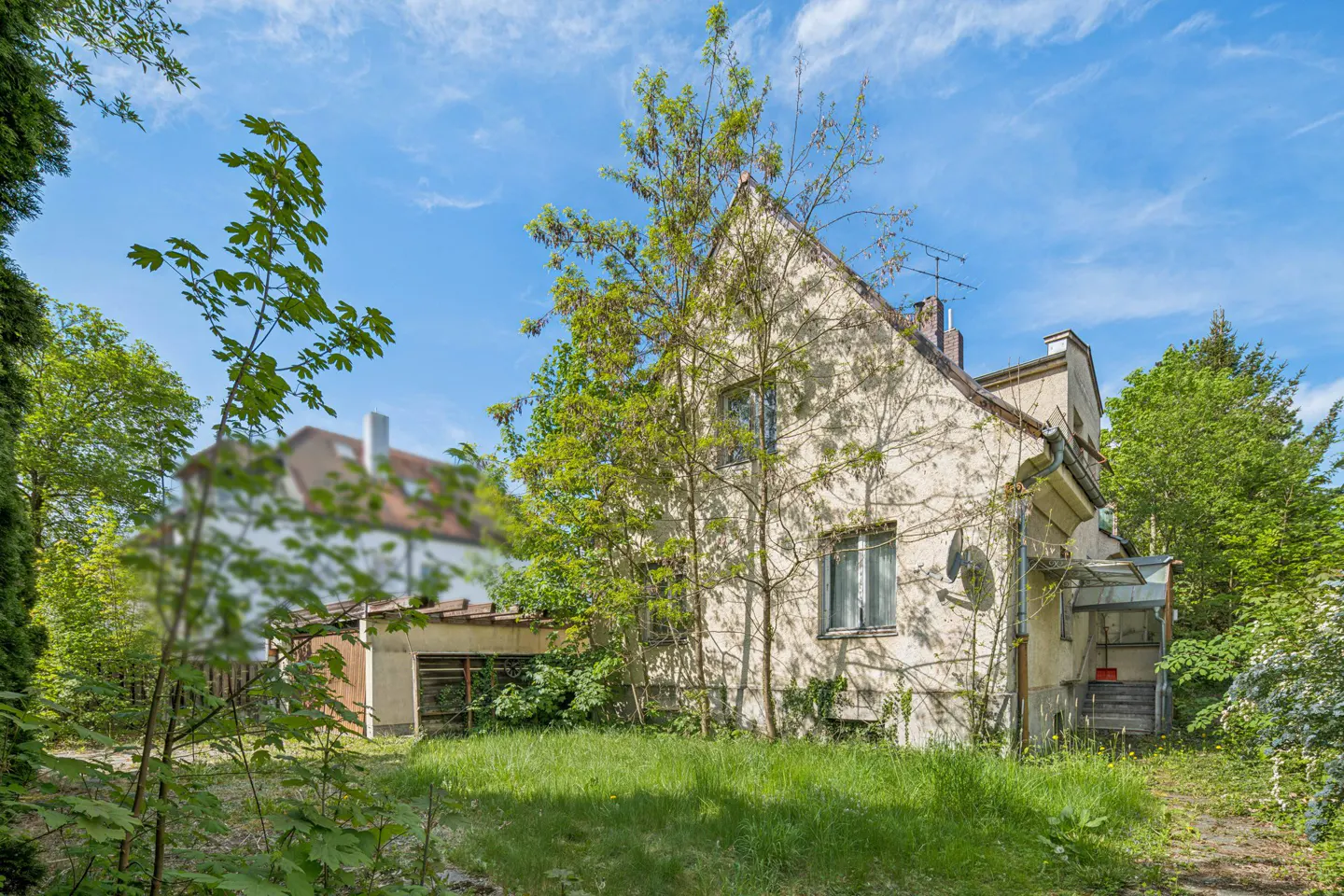 Exterior view of a two-story beige house with a gable roof, surrounded by green trees and grass under a blue sky.