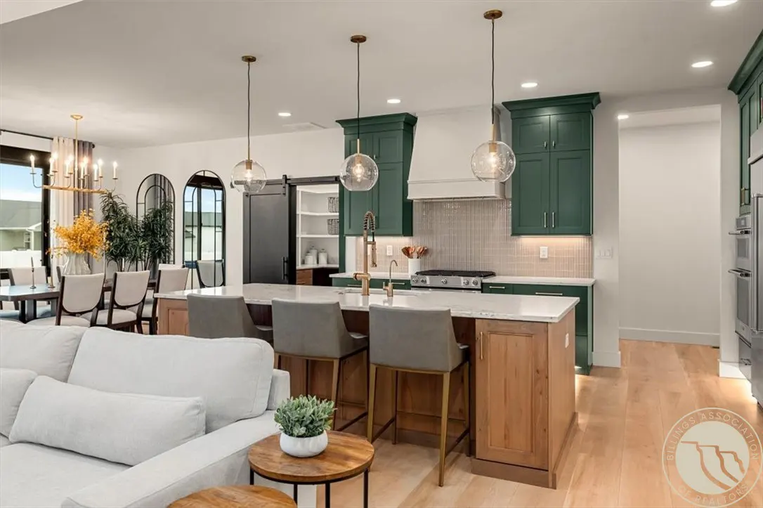 Open-concept kitchen with green cabinets, a wood island with gray stools, and globe pendant lights. A white sofa is in the foreground.