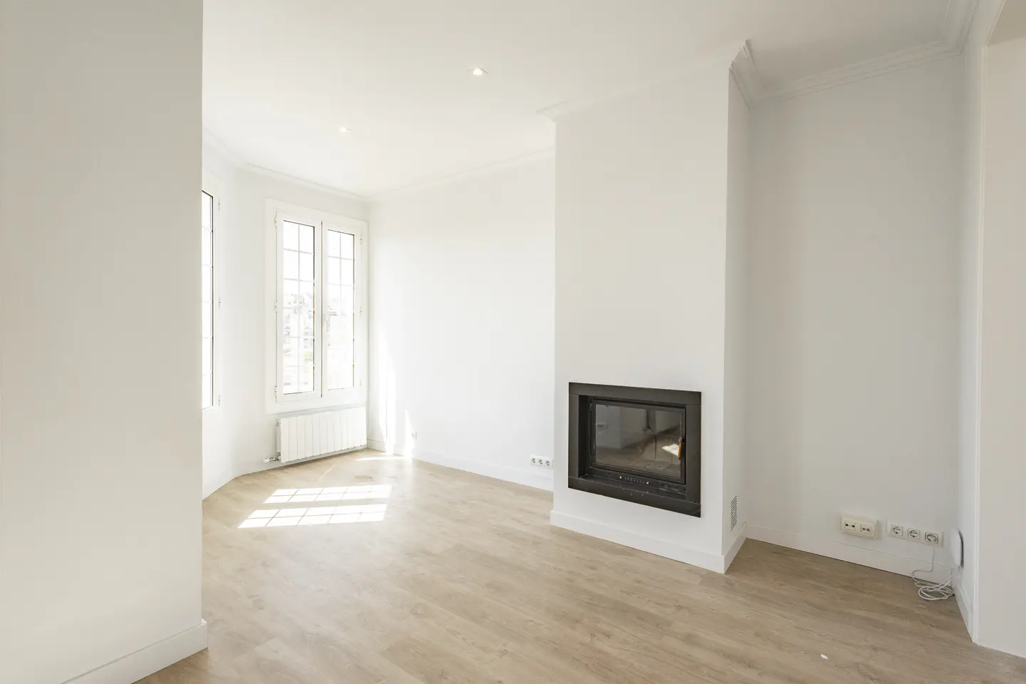 Bright, empty room with light wood floors, white walls, and a black fireplace. Windows let in sunlight.
