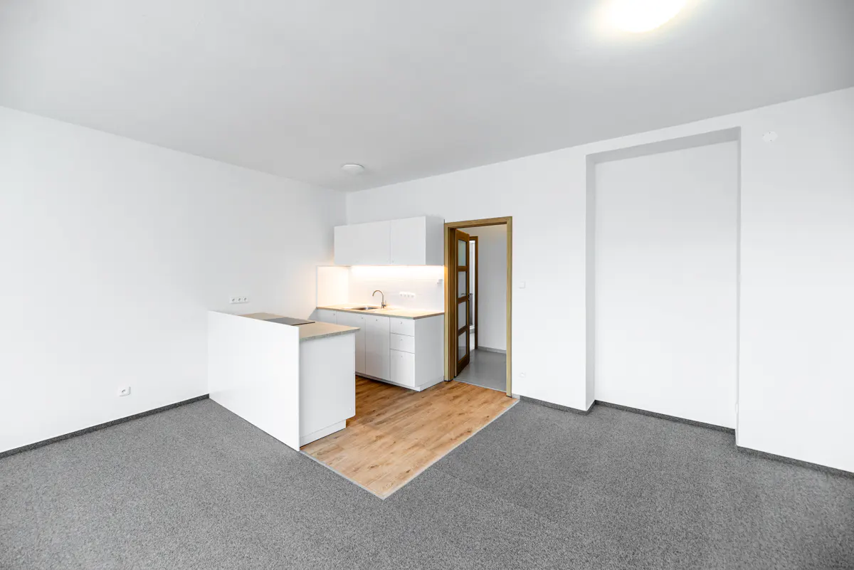 Bright, modern apartment with white walls, gray carpet, and a small kitchen area with white cabinets and wood flooring. A doorway is visible.