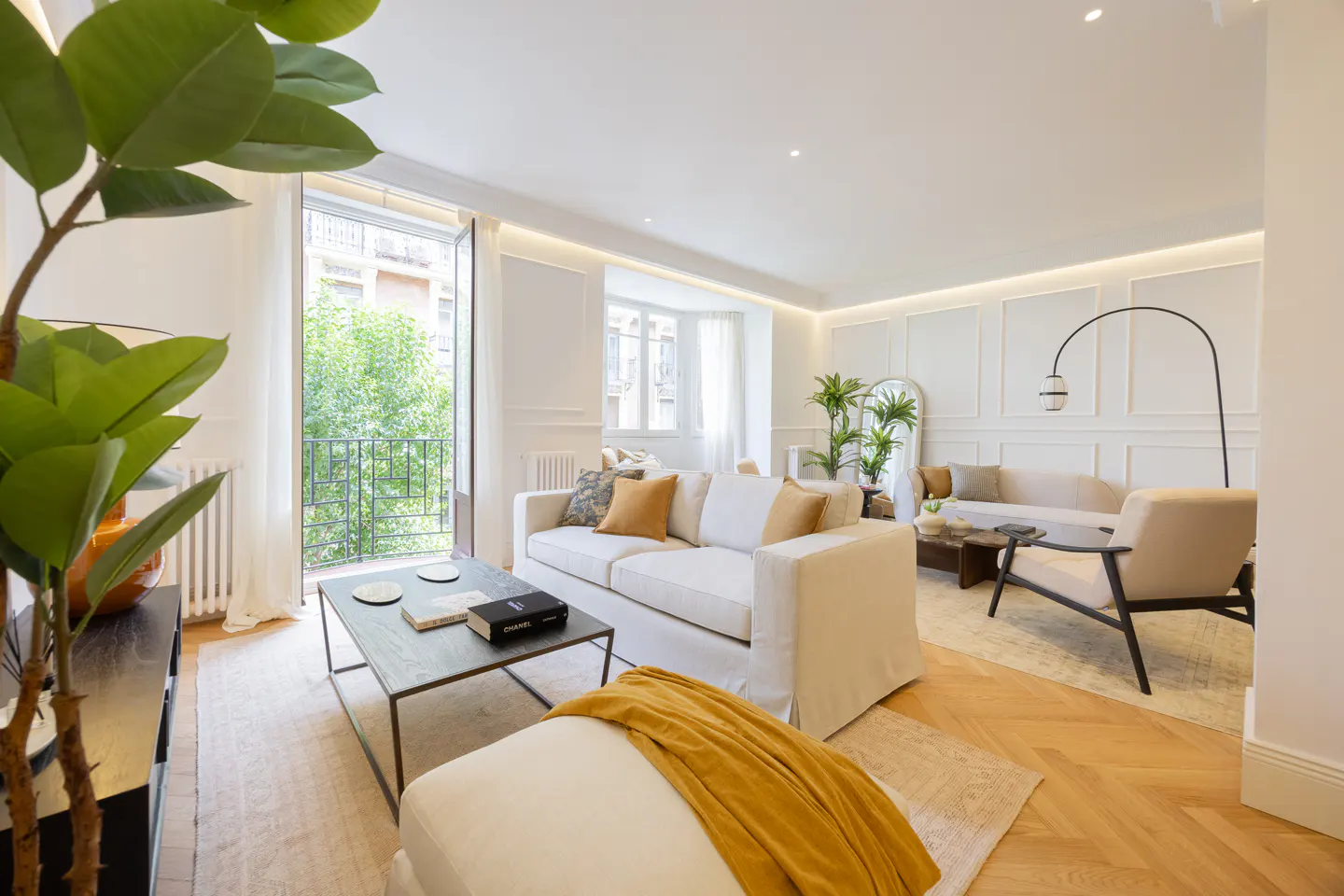Bright, modern living room with white sofas, a mustard throw, herringbone wood floors, and a balcony with green trees.