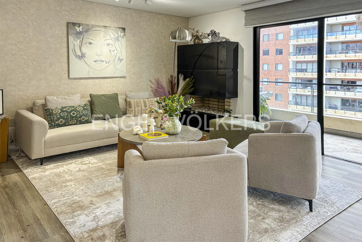 Modern living room with beige sofa, two armchairs, round coffee table, and large wall art, overlooking a city building.