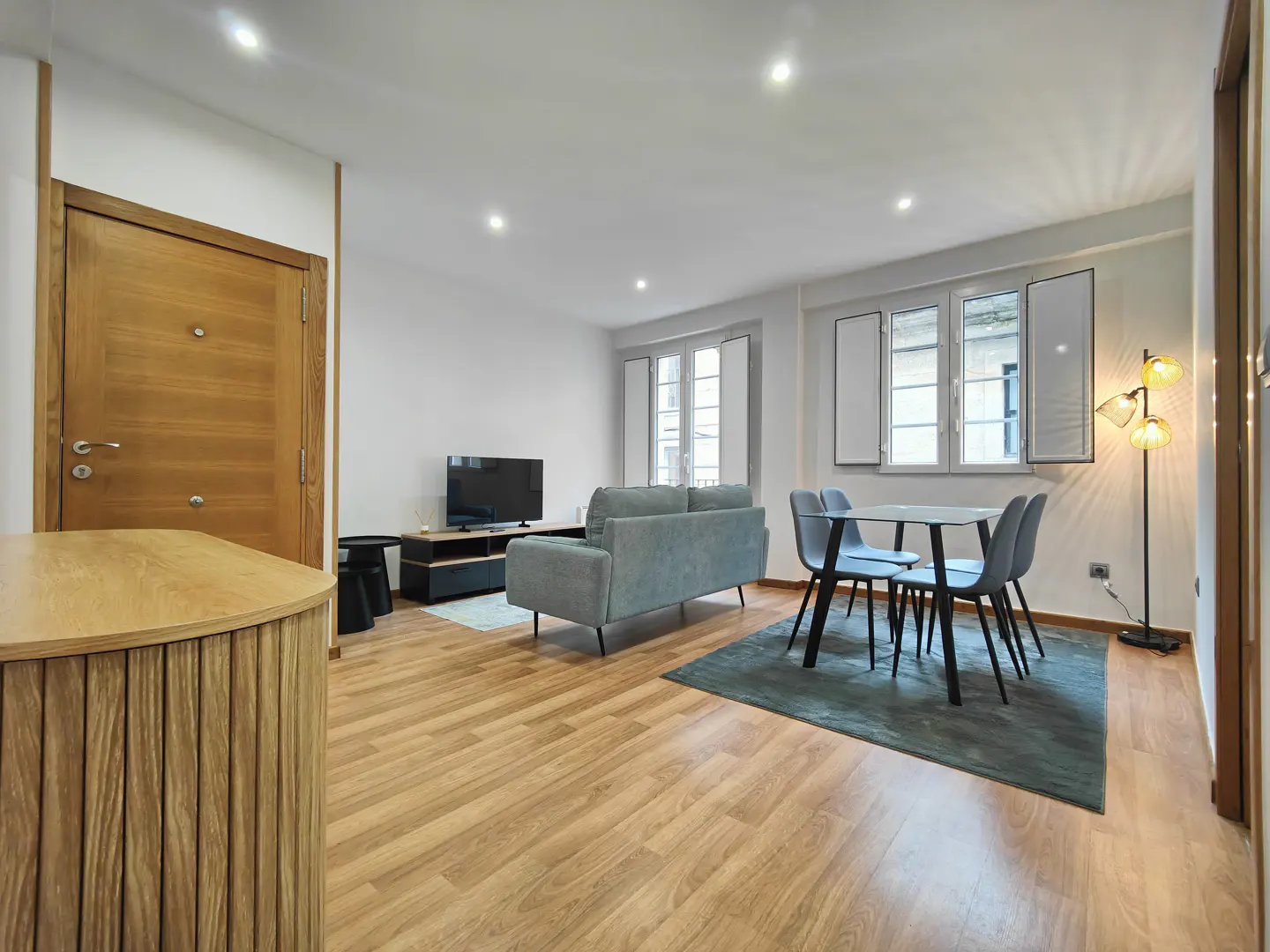 Bright apartment interior with wood floors, a gray sofa, a dining table with four chairs, and a TV. Natural light from windows.