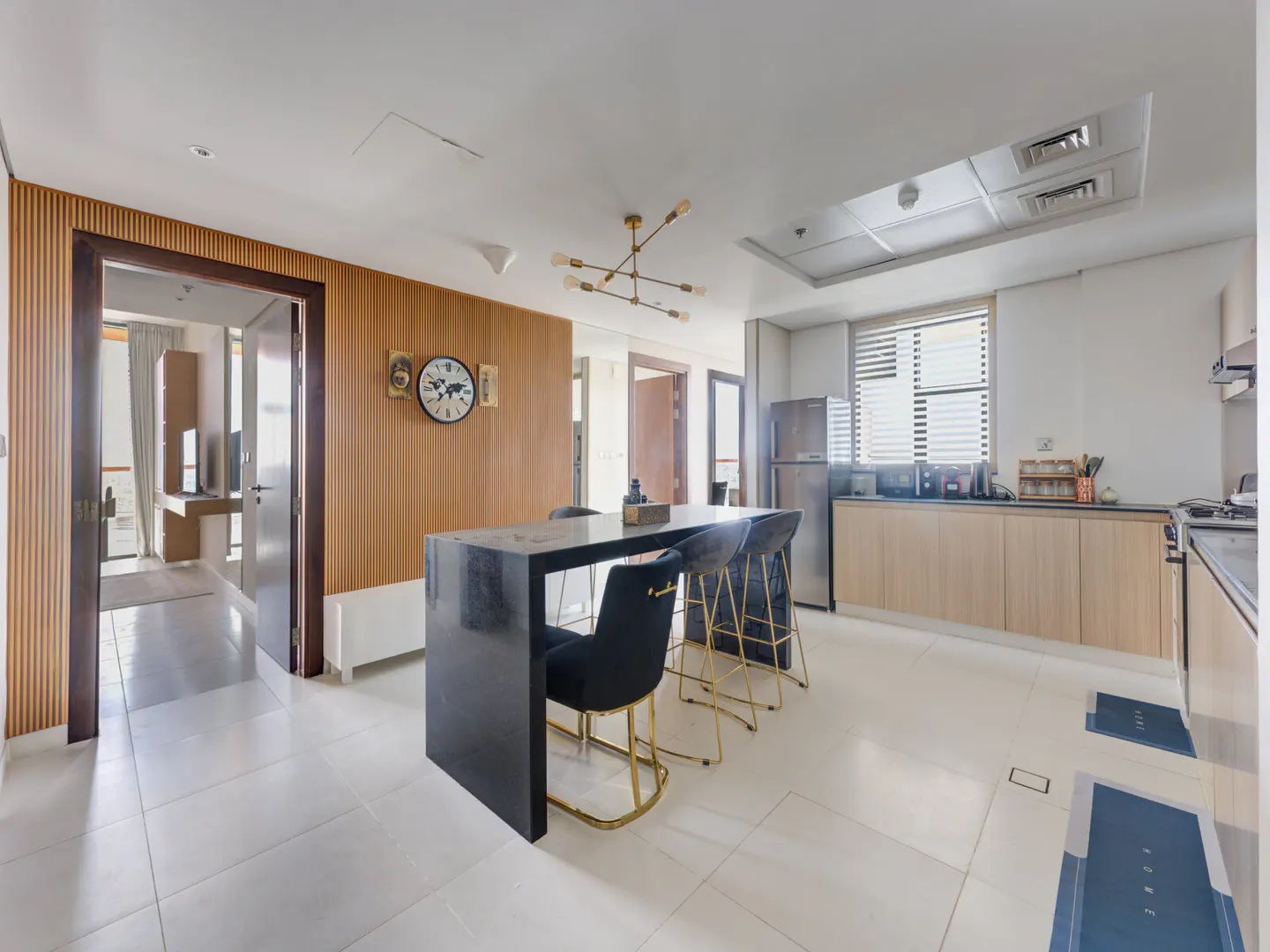 A modern apartment interior with a black kitchen island, gold chairs, and light wood cabinets. A clock hangs on a wood-paneled wall.