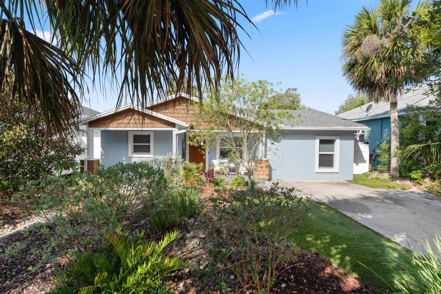 A light blue single-story house with a brown roof is surrounded by lush green landscaping and palm trees.