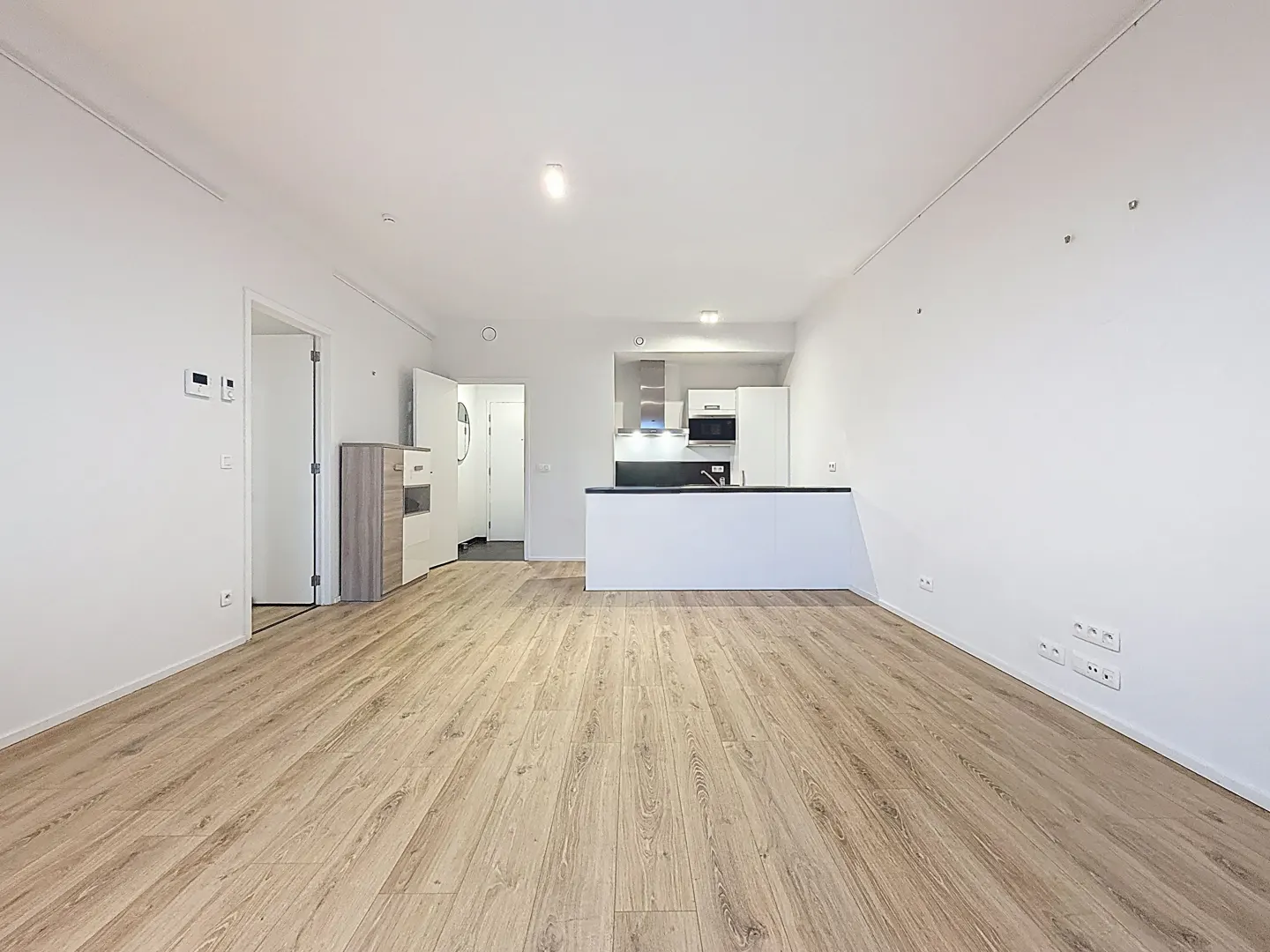 Bright, open-plan living area with light wood flooring, white walls, and a modern kitchen island in the background.
