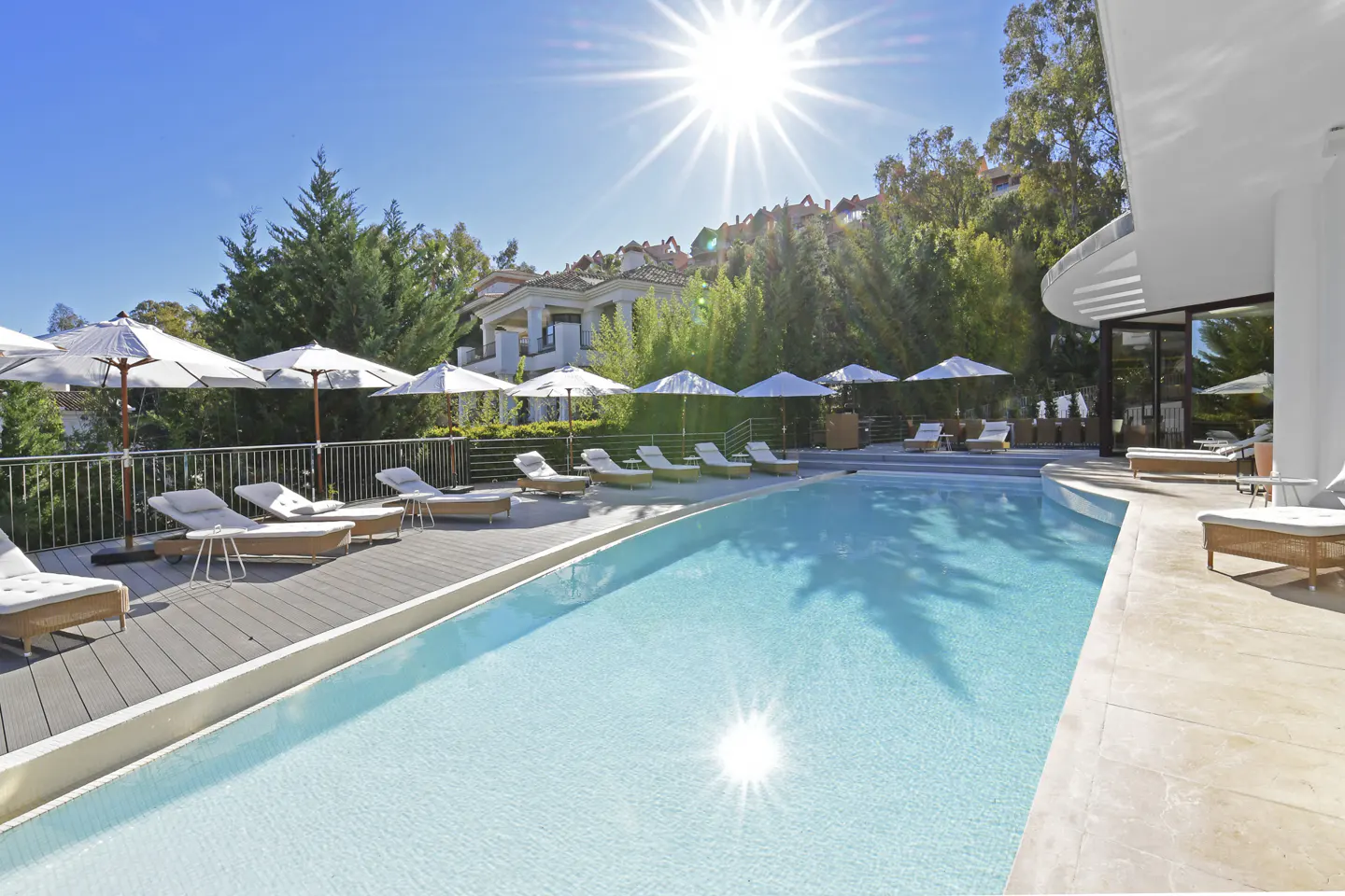 A bright, sunny day illuminates a luxurious pool area with lounge chairs and umbrellas. Trees and a building are in the background.