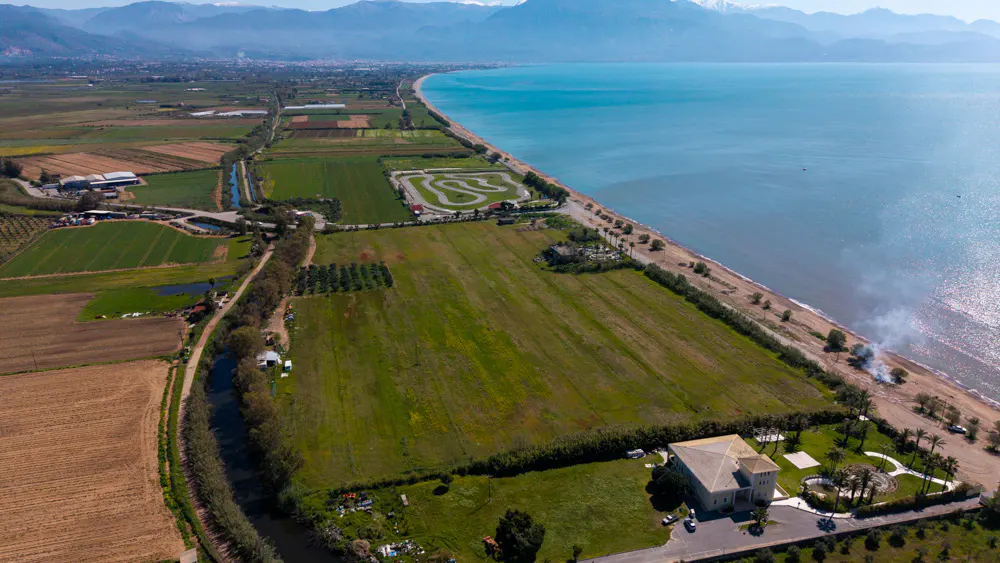 Aerial view of a coastal landscape with green fields, a go-kart track, a sandy beach, and turquoise sea, backed by mountains.