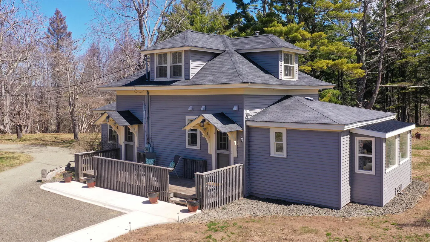 Two-story gray house with a dark gray roof, a wooden deck, and a gravel driveway surrounded by trees.
