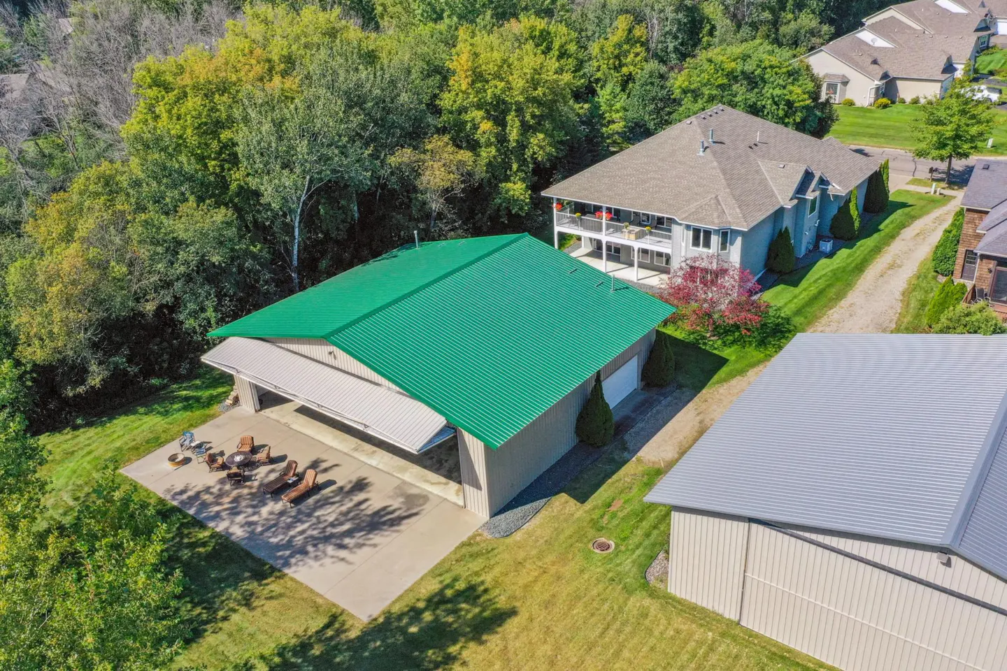 Aerial view of a tan building with a green roof, surrounded by trees and other houses. Patio furniture sits on the concrete pad in front.