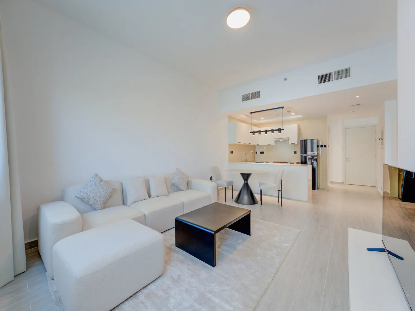 Bright, open-concept apartment with a white sectional sofa, black coffee table, and kitchen with a round table and chairs.