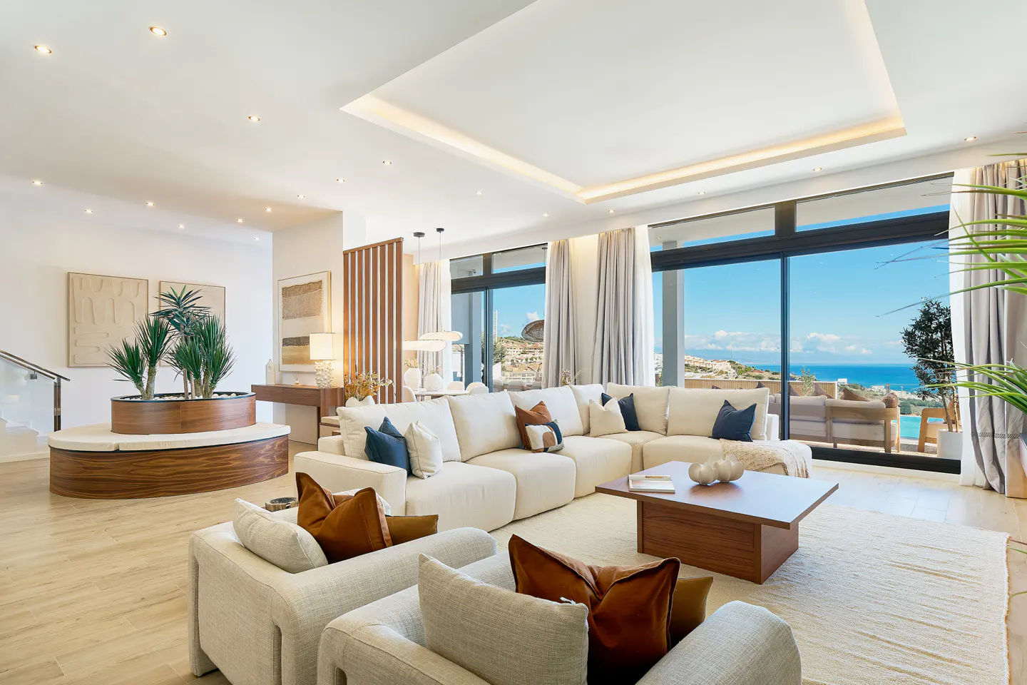 Bright, modern living room with a large white sectional sofa, wood coffee table, and ocean view through large windows. Neutral tones and natural light.