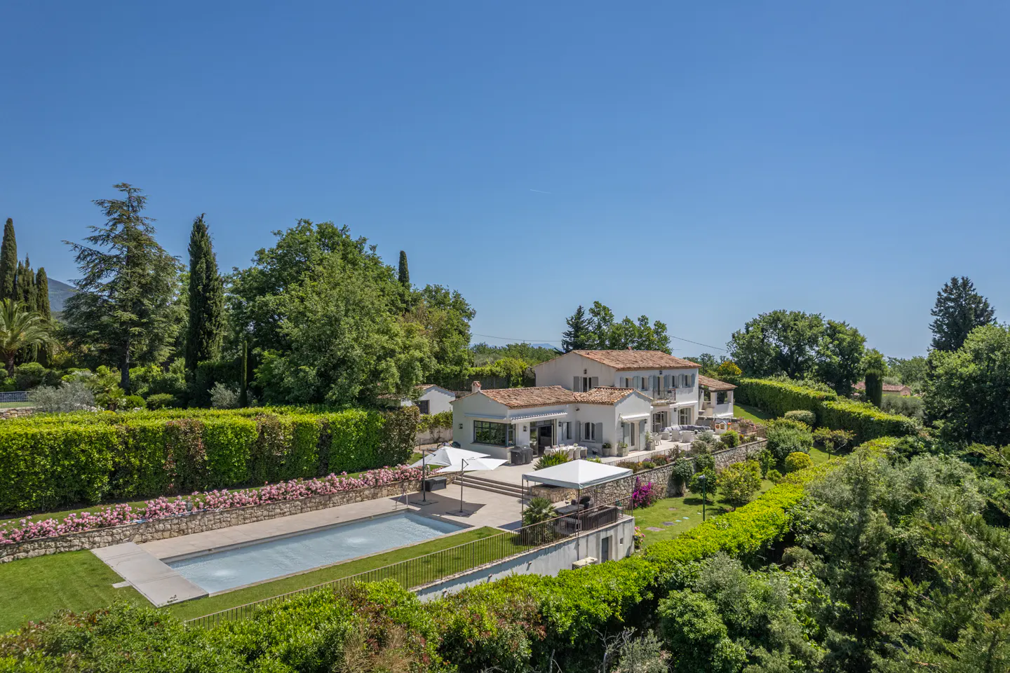 Aerial view of a white, tile-roofed house with a pool, surrounded by green trees and hedges under a clear blue sky.