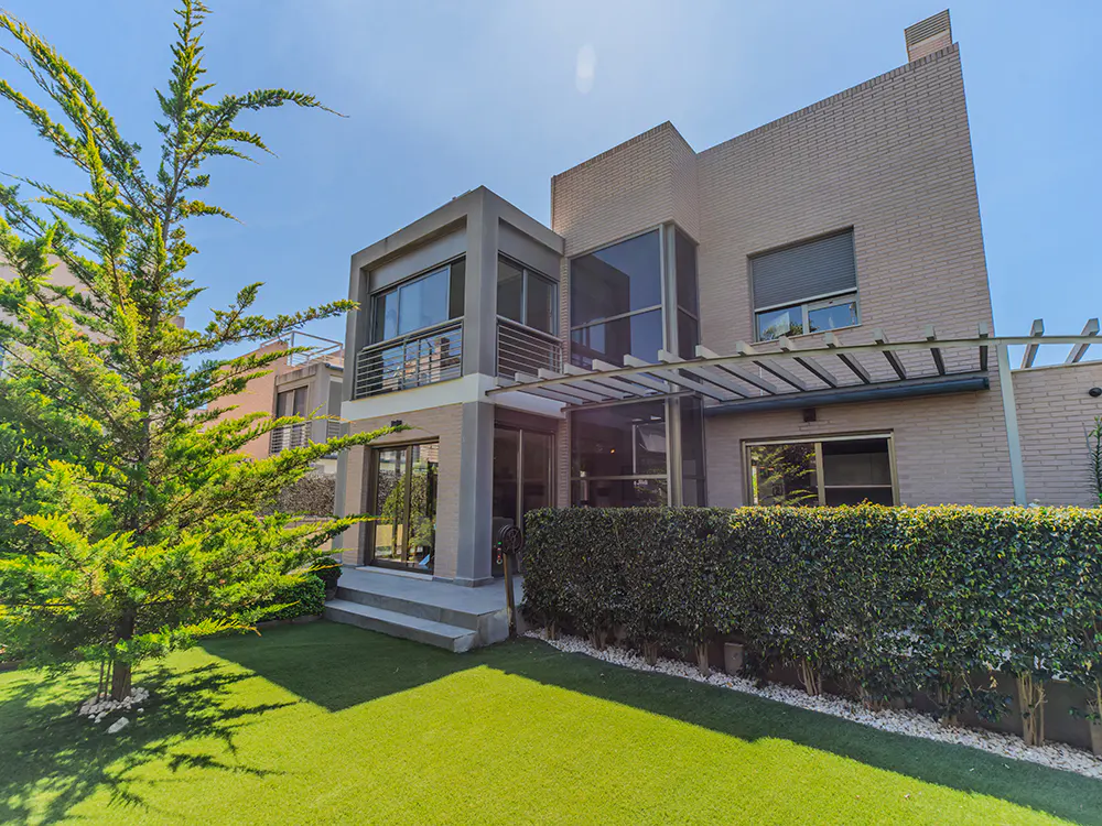 Modern two-story house with a green lawn, a tall tree, and a trimmed hedge under a sunny blue sky.