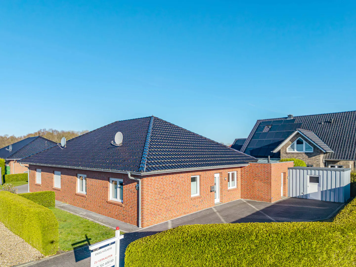A red brick house with a dark roof and white trim, surrounded by green hedges, under a clear blue sky. A "For Sale" sign is in the front yard.