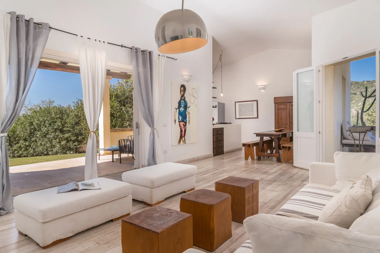 Bright living room with white sofa, ottomans, and wooden stools. Open doors lead to a patio with green foliage. Artwork and dining set in the background.