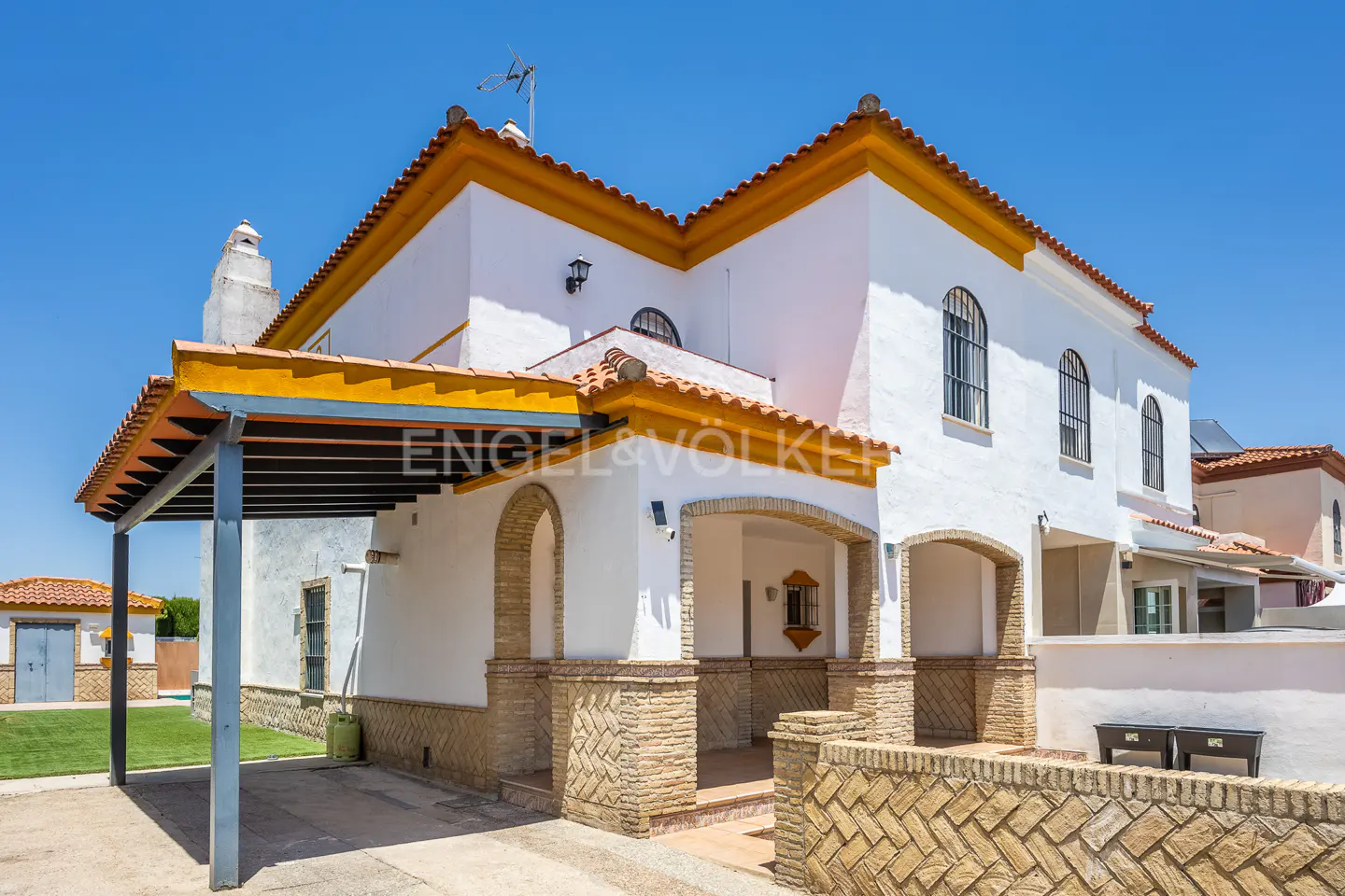 Exterior of a two-story white house with orange trim and a tile roof under a clear blue sky. A covered porch with stone arches is visible.