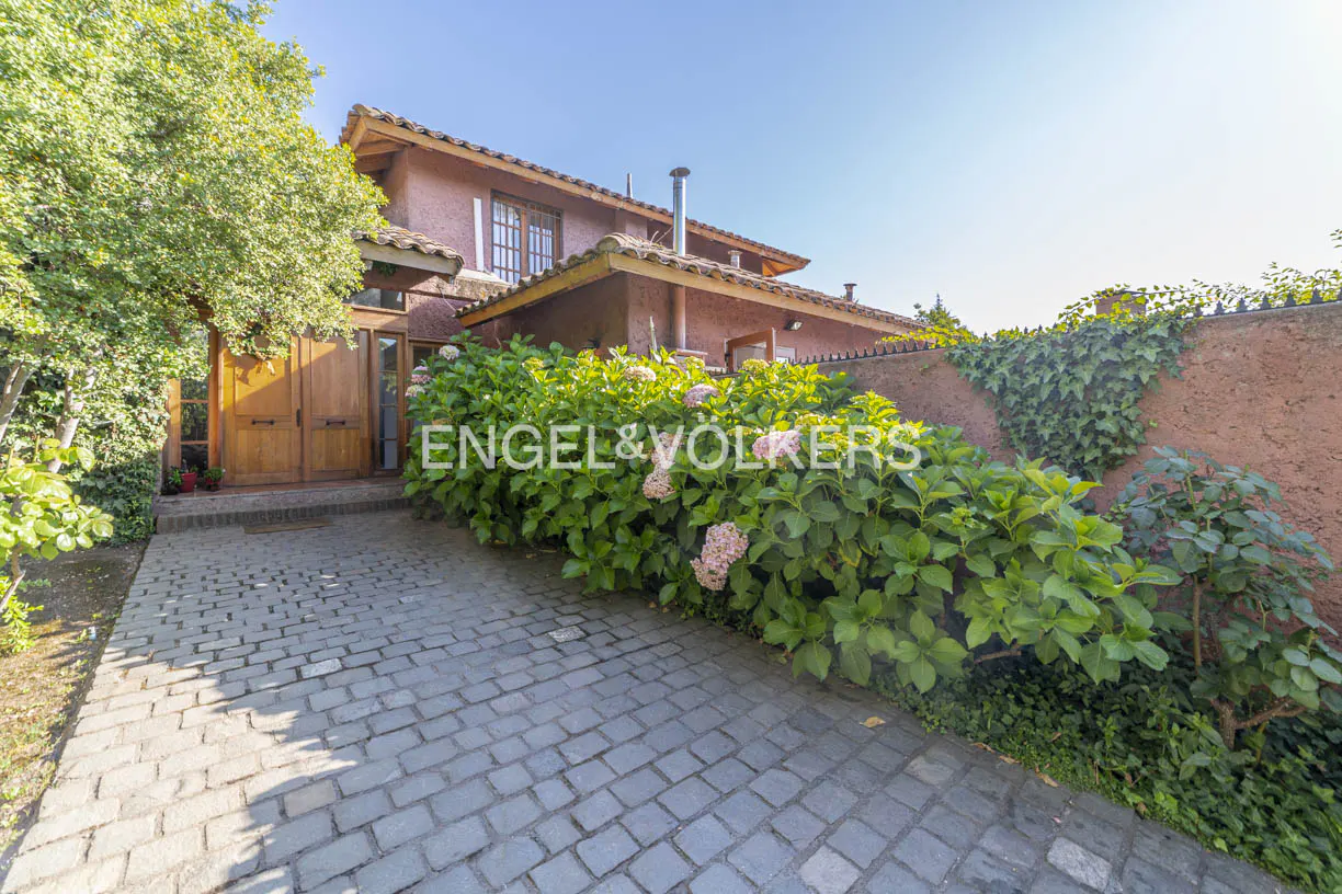 Exterior view of a two-story pink house with a stone driveway and lush green landscaping. The Engel & Völkers logo is superimposed on the image.