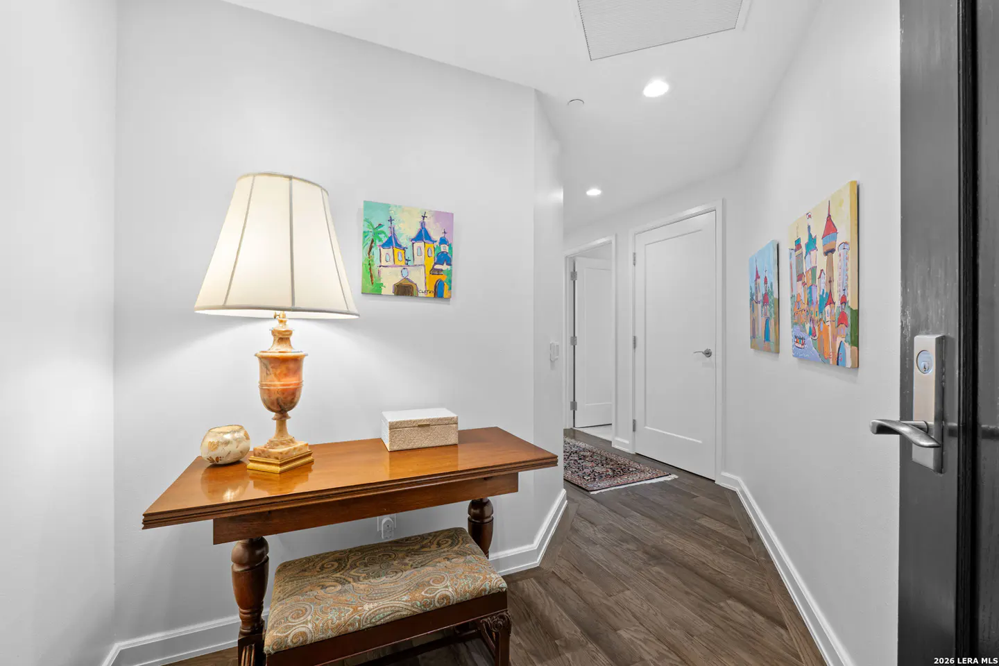 Hallway with a wooden table, lamp, and bench. Paintings adorn the white walls, and a dark wood floor leads to white doors.