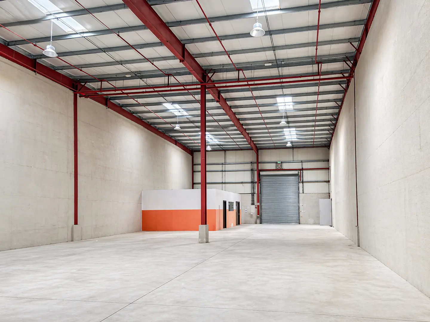 Empty warehouse interior with concrete floor, white walls, and red steel support beams. Skylights and hanging lights illuminate the space. A gray roll-up door is at the back.