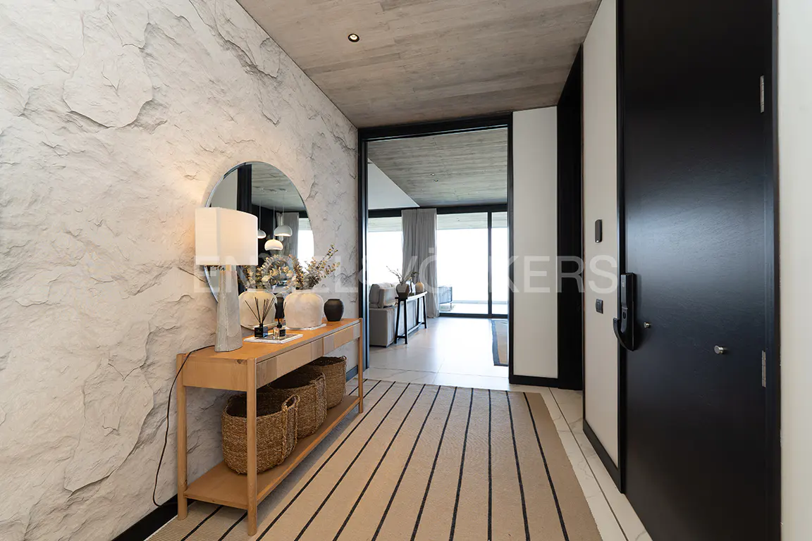 A modern entryway with a stone wall, wood console table, round mirror, and black door. A striped rug leads to a bright living space.