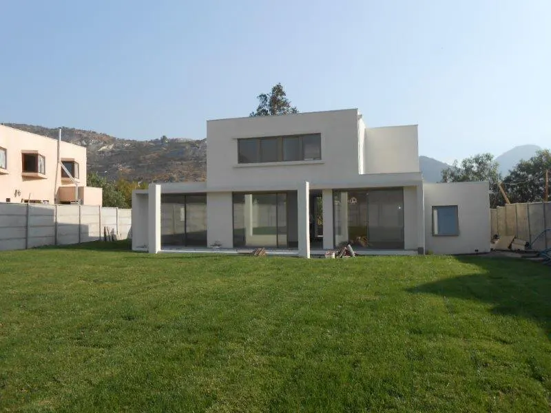 A modern white house with large windows sits on a green lawn, under a clear blue sky. A concrete fence surrounds the property.