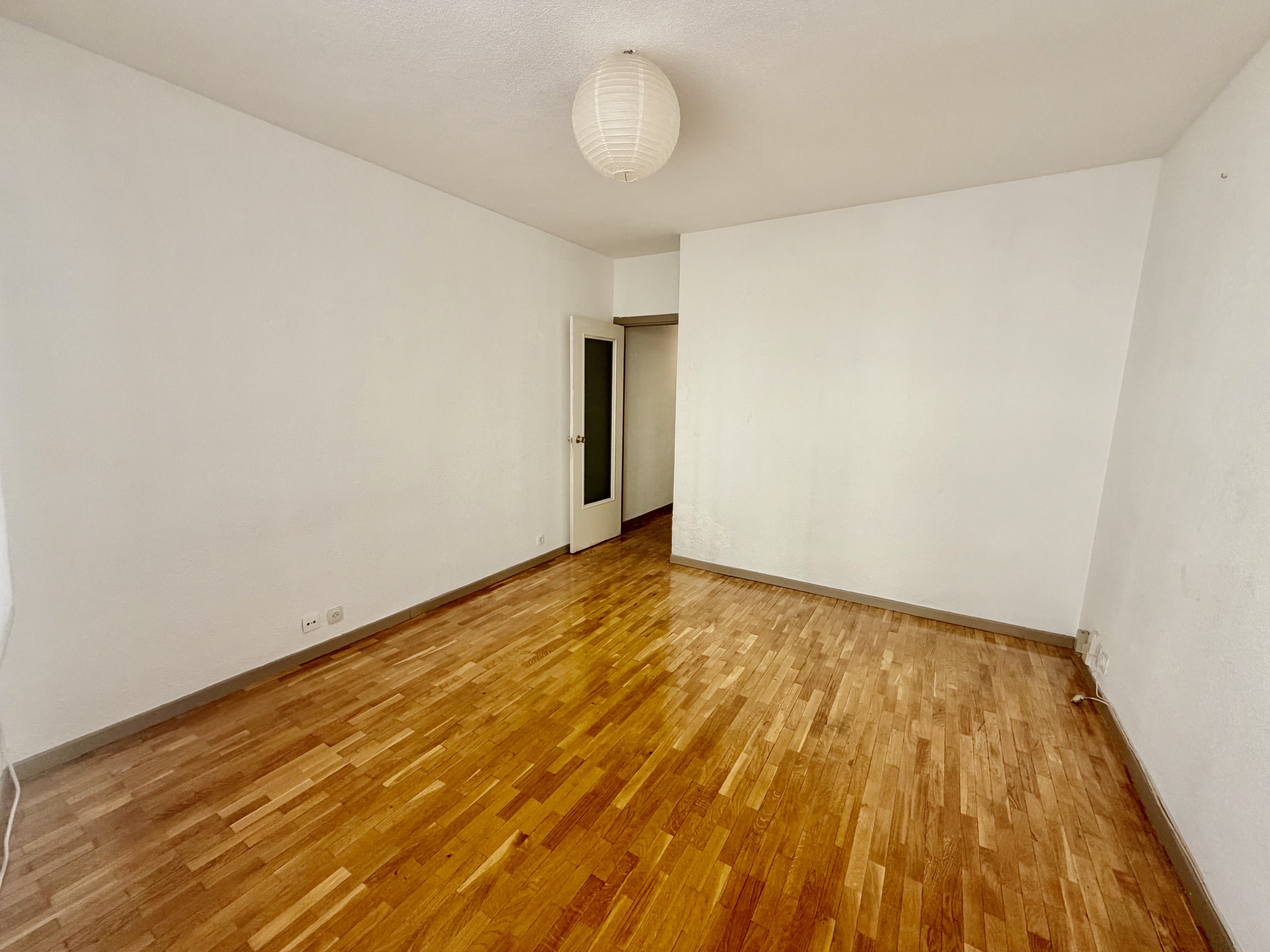 Empty room with white walls, polished light wood flooring, a round paper ceiling lamp, and a white door leading to a hallway.