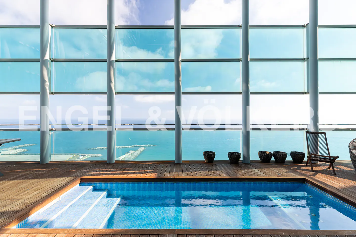 Indoor pool with blue tiles and wooden deck, overlooking the ocean through a large window. A chair and pots are on the deck.