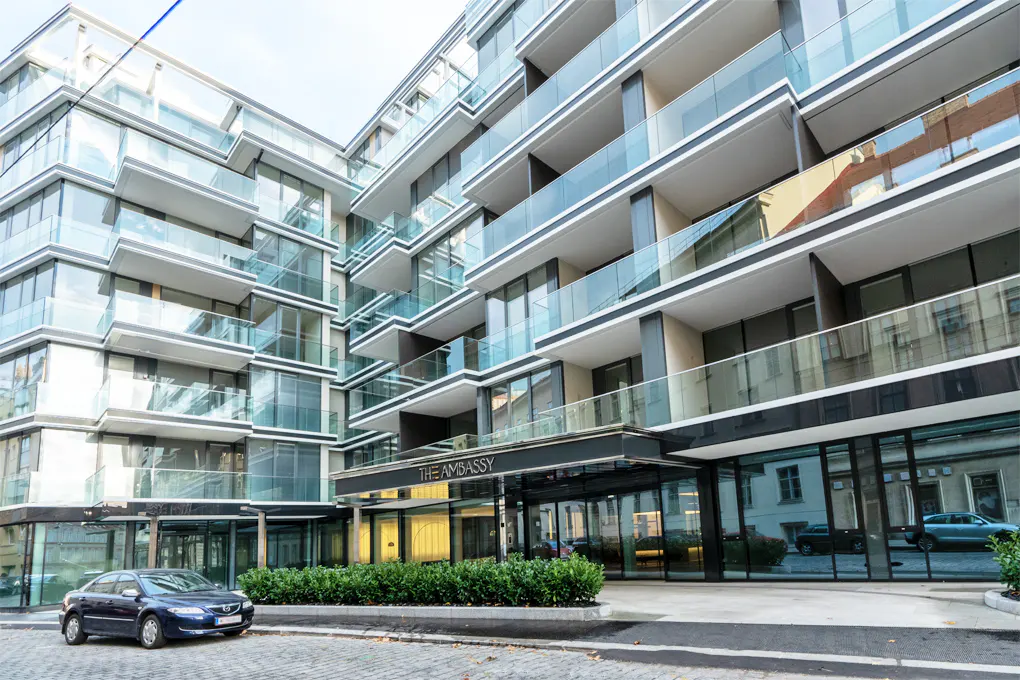 Modern apartment building, "The Embassy," with glass balconies. A blue car is parked in front.