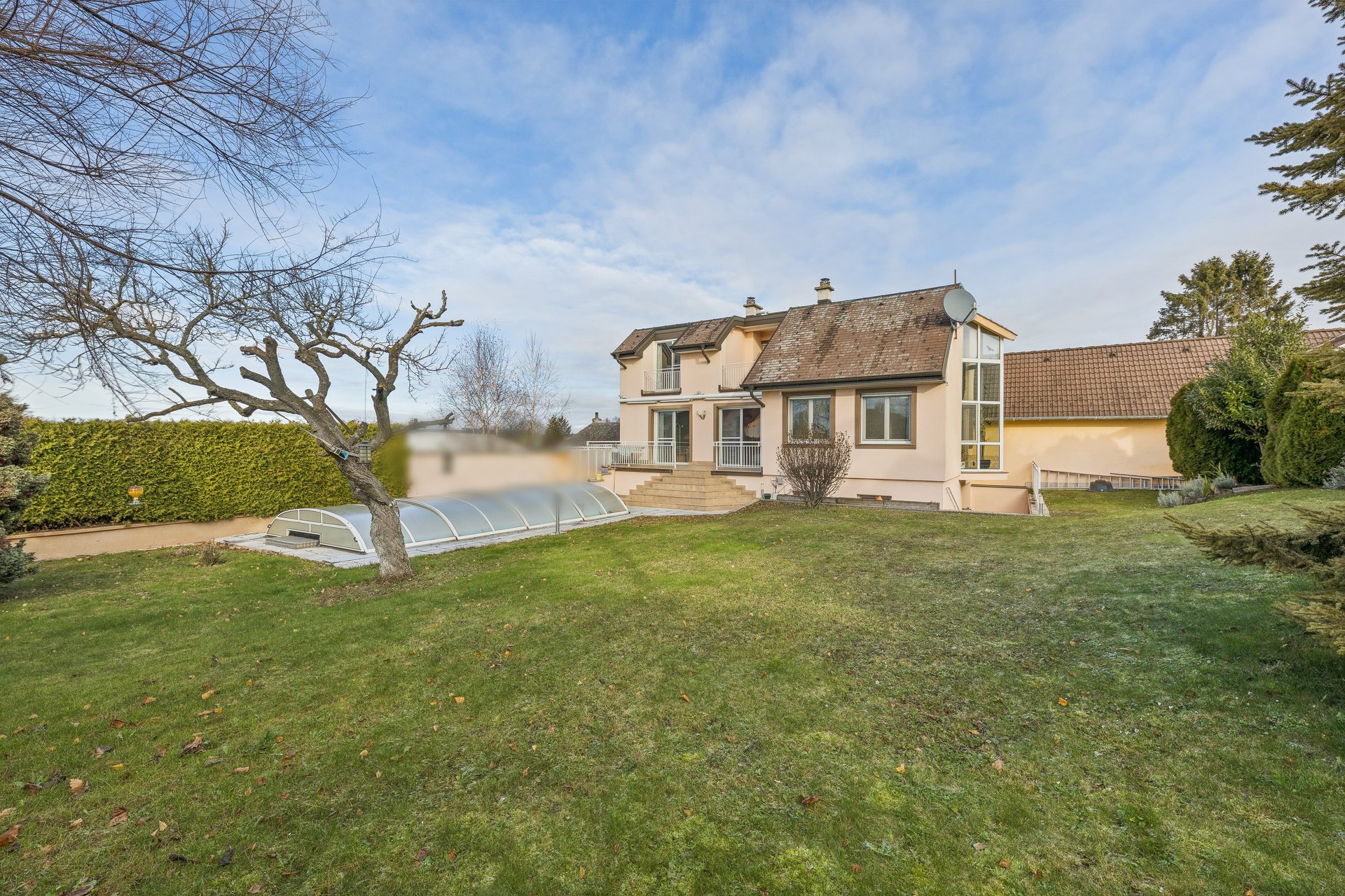 Backyard of a beige two-story house with a covered swimming pool, green lawn, and bare trees under a blue sky.