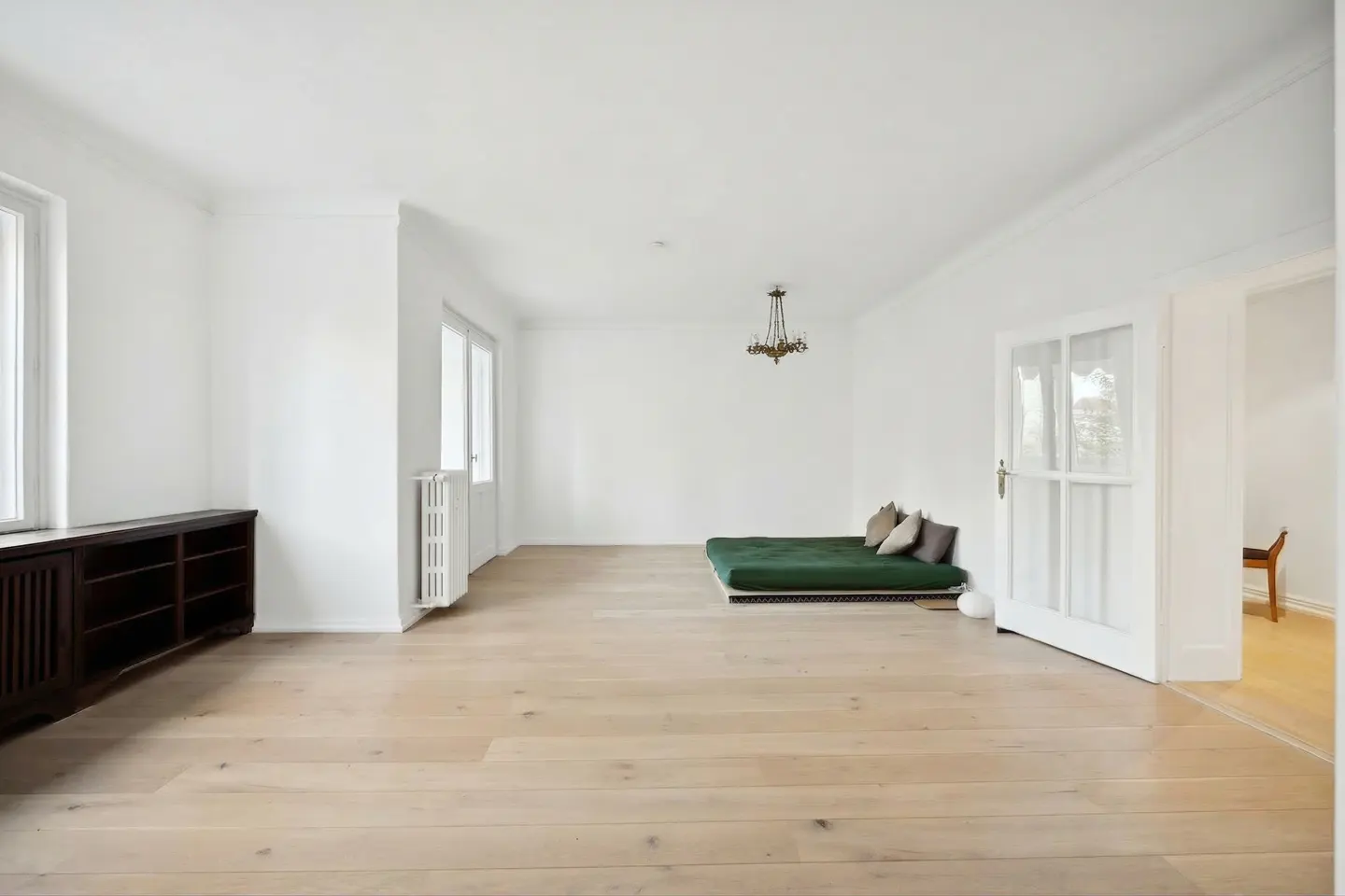 Bright, empty room with white walls, wood floors, and a green futon with pillows. A dark wood cabinet sits near a window. A chandelier hangs from the ceiling.