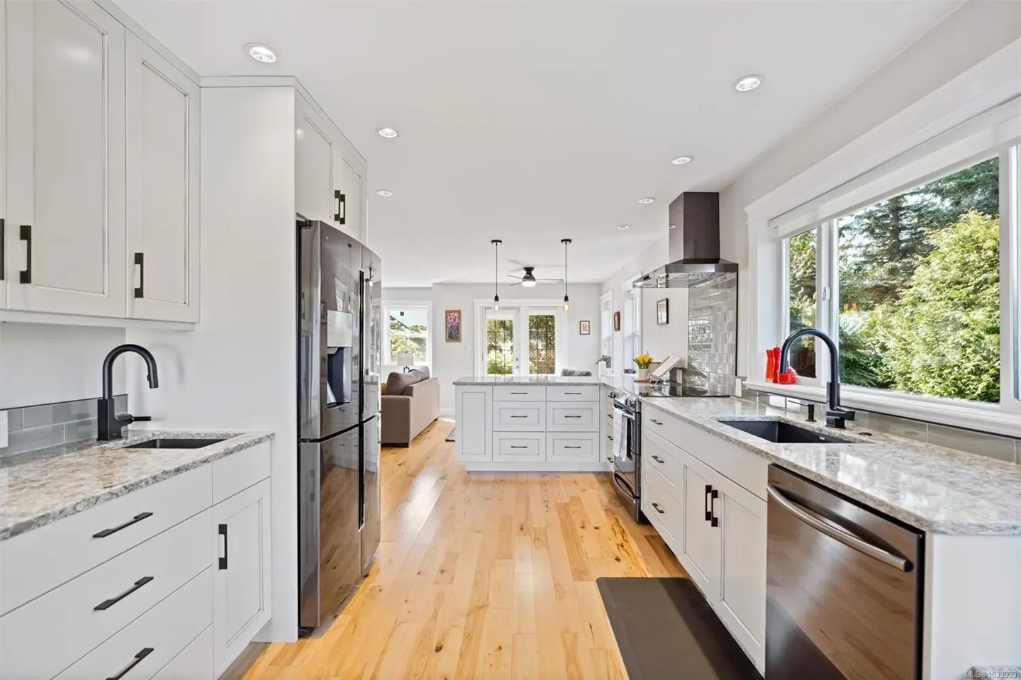 Bright, open kitchen with white cabinets, granite countertops, and stainless steel appliances. Hardwood floors lead to a living area.