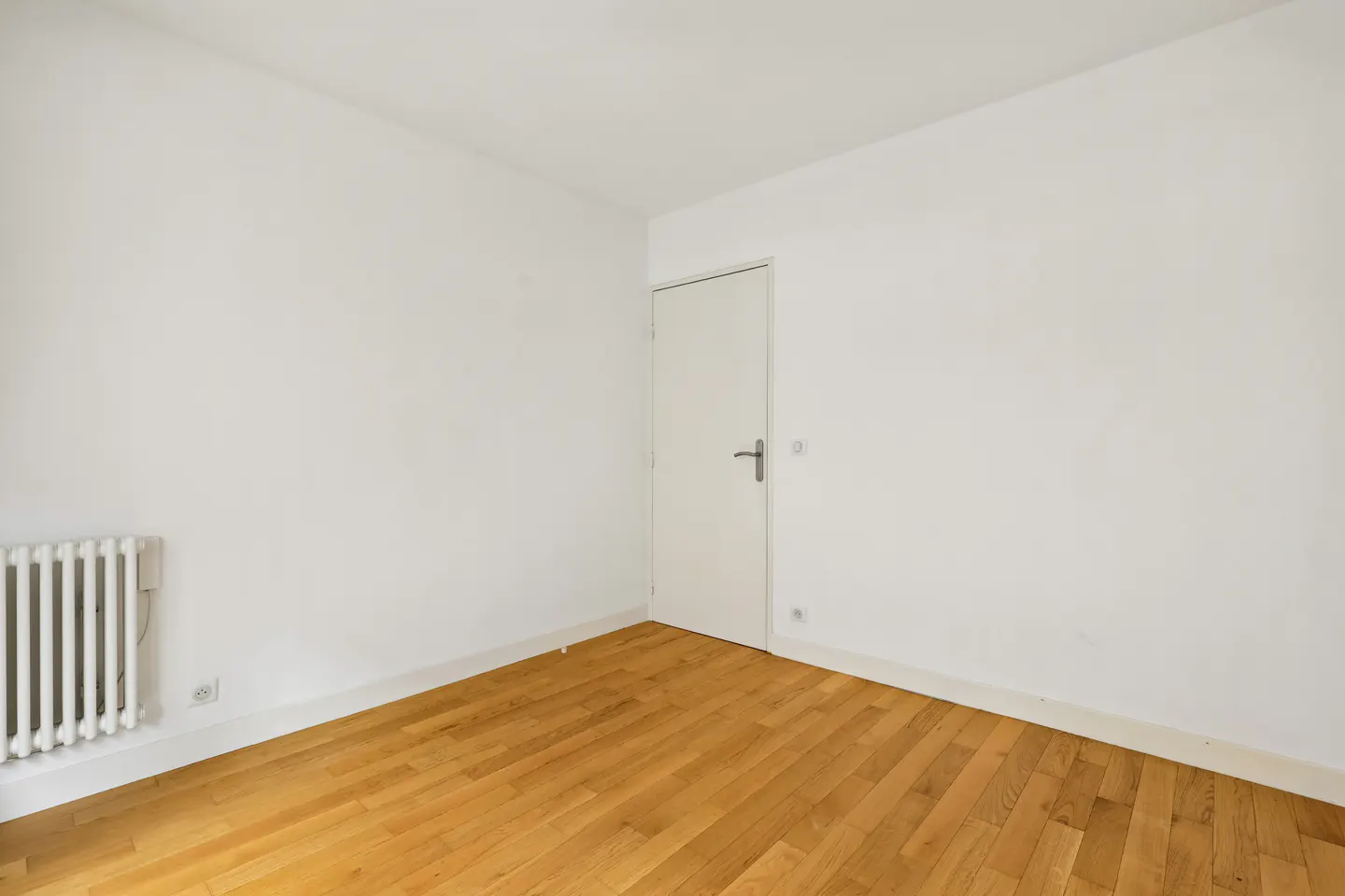Empty room with white walls, a light wood floor, a white radiator, and a white door.