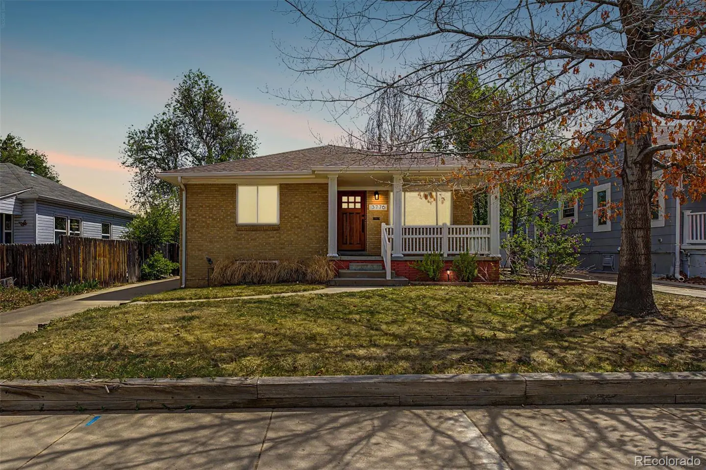 Tan brick house with a white porch and a brown front door, set against a blue sky.