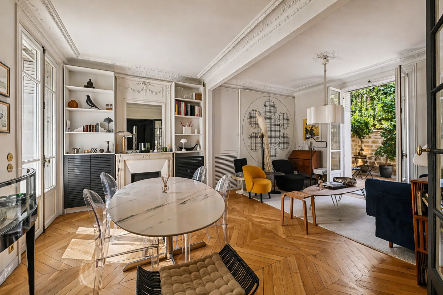Bright, open-concept living and dining room with herringbone wood floors. A marble table with clear chairs sits near built-in shelves. A garden is visible through open doors.