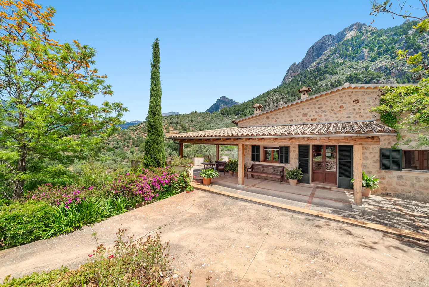 Stone house with a tiled roof, a covered porch, and a mountain backdrop on a sunny day.