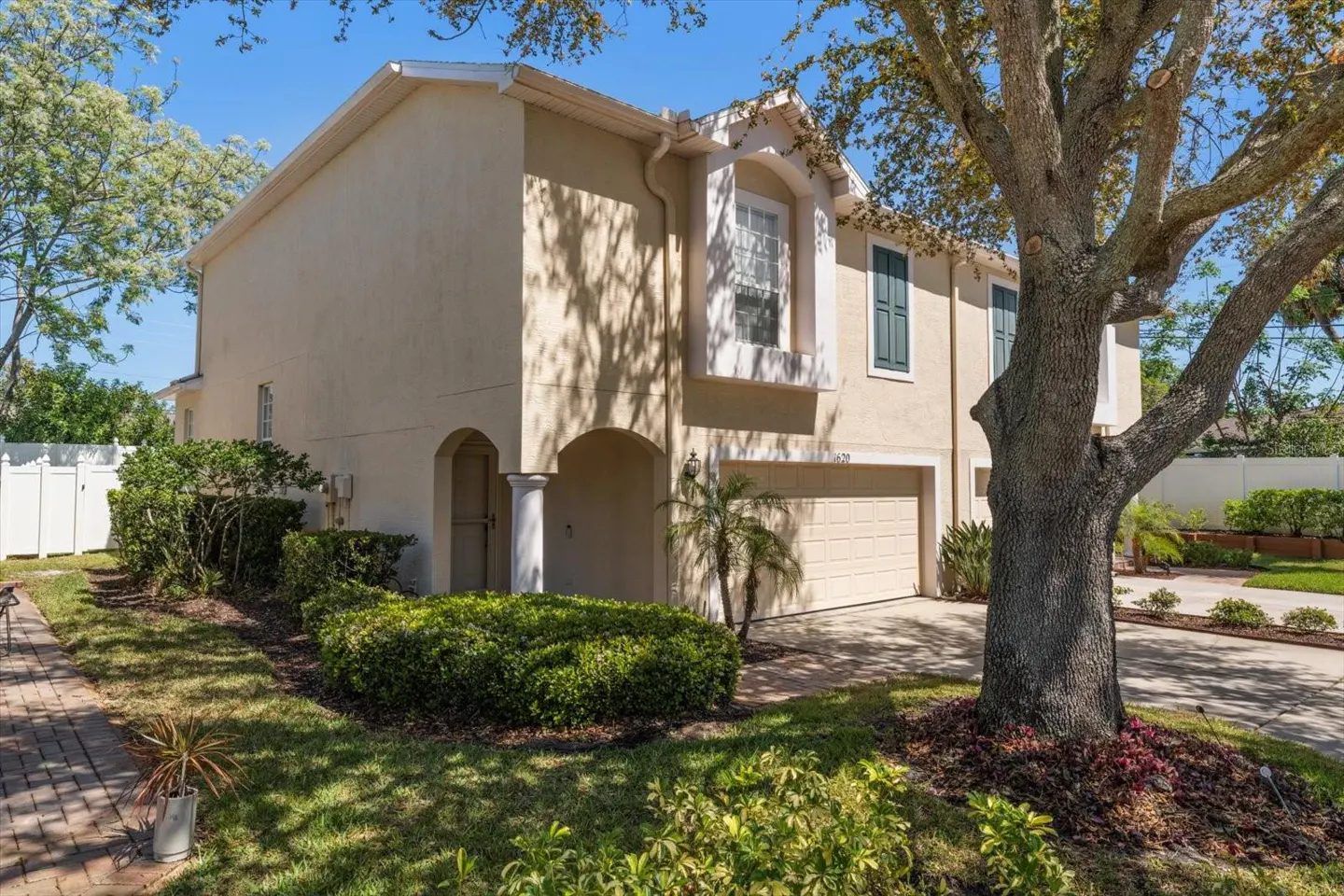 Beige two-story house with a two-car garage, green shutters, and a large tree in the front yard.
