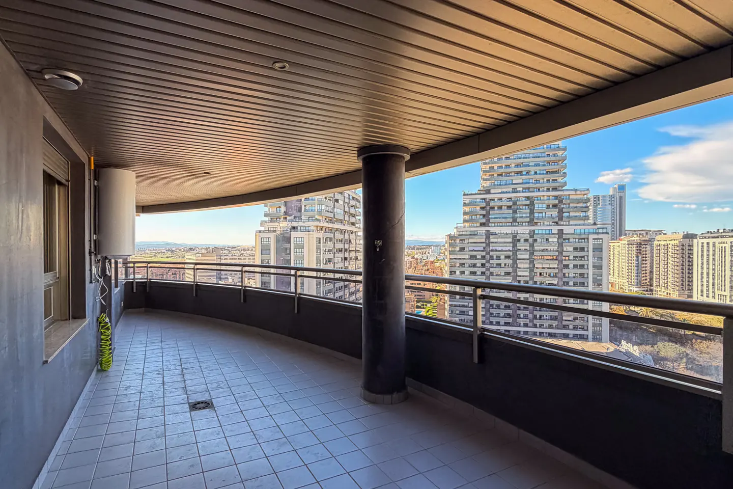 Wide balcony with gray tile floor, dark railing, and a view of city buildings under a blue sky.