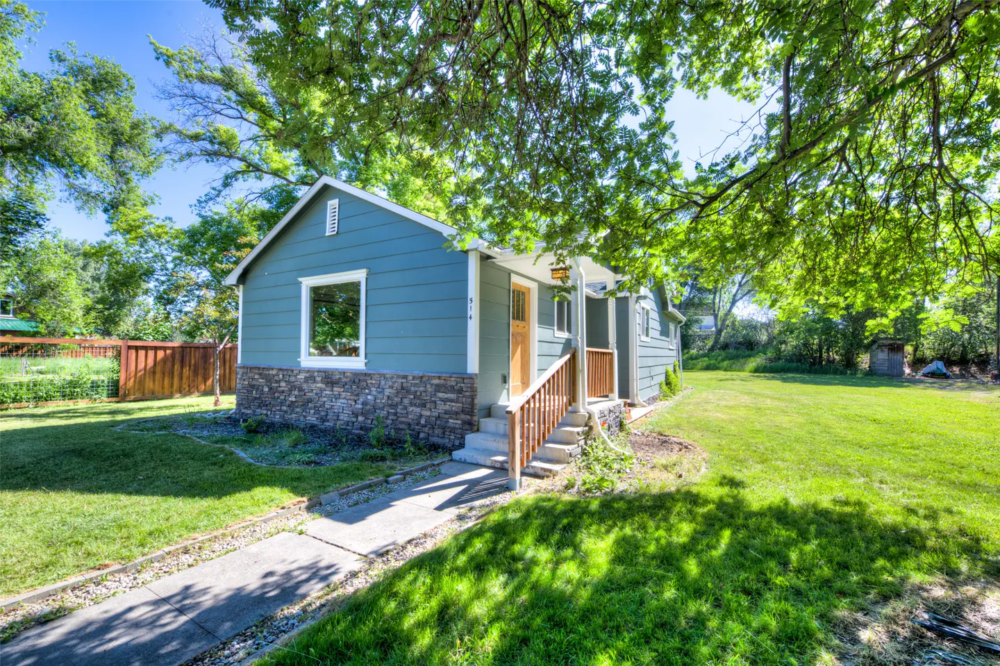 Blue house with stone siding, wooden front porch, and a concrete walkway on a sunny, tree-shaded lawn.