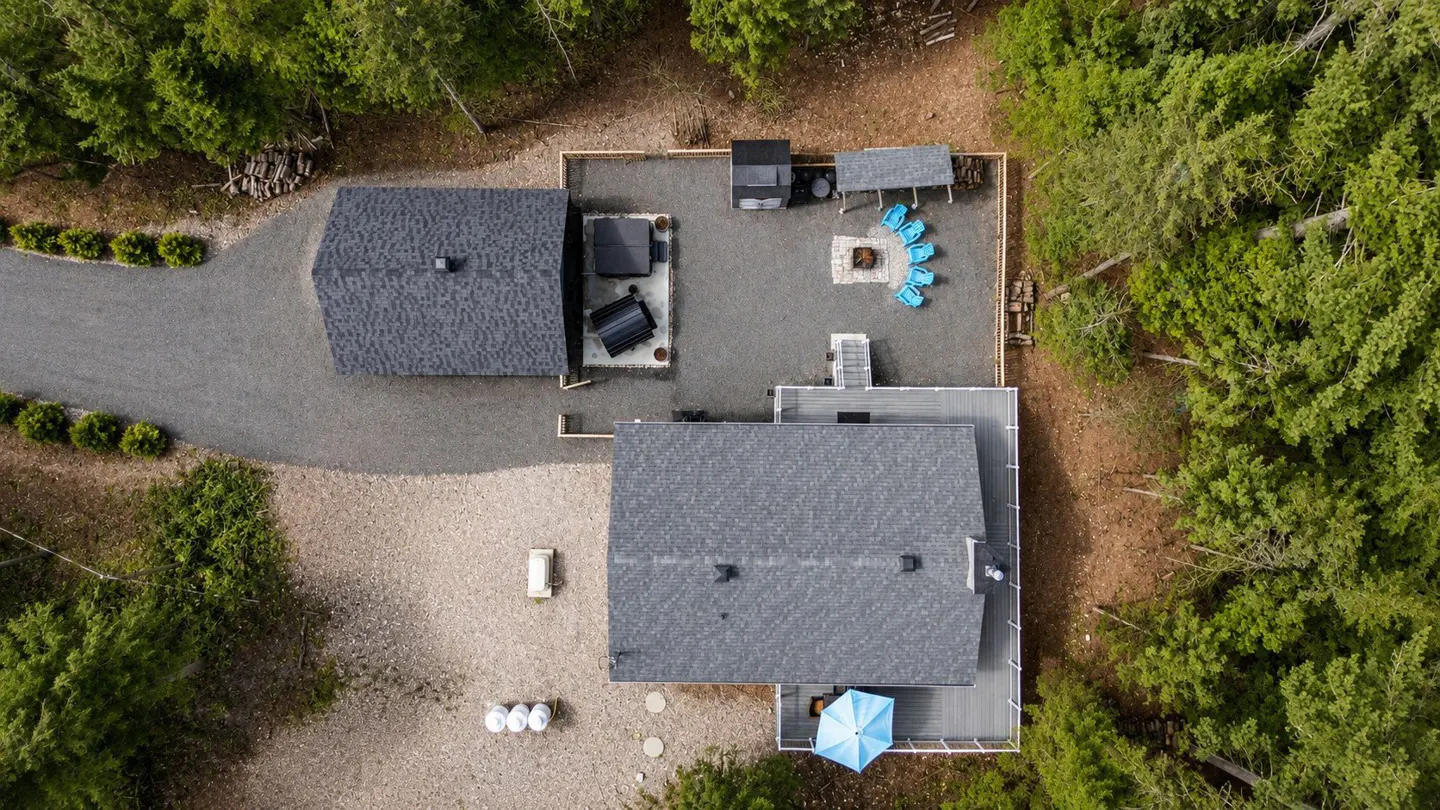 Aerial view of a gray-roofed house with a deck, blue umbrella, and a gravel yard with a fire pit surrounded by blue chairs. Trees surround the property.