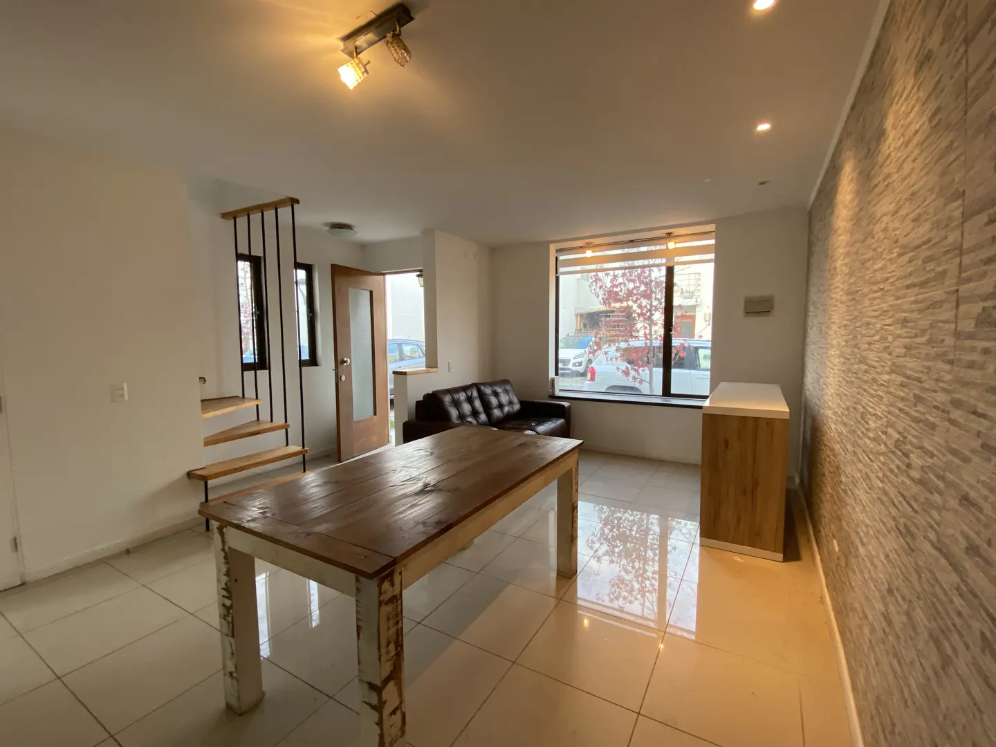 A bright living room with a wooden table, leather sofa, and a staircase. A large window lets in natural light.