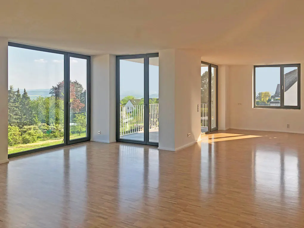 Bright, empty room with light wood floors, white walls, and large windows. Balcony access and green landscape views.