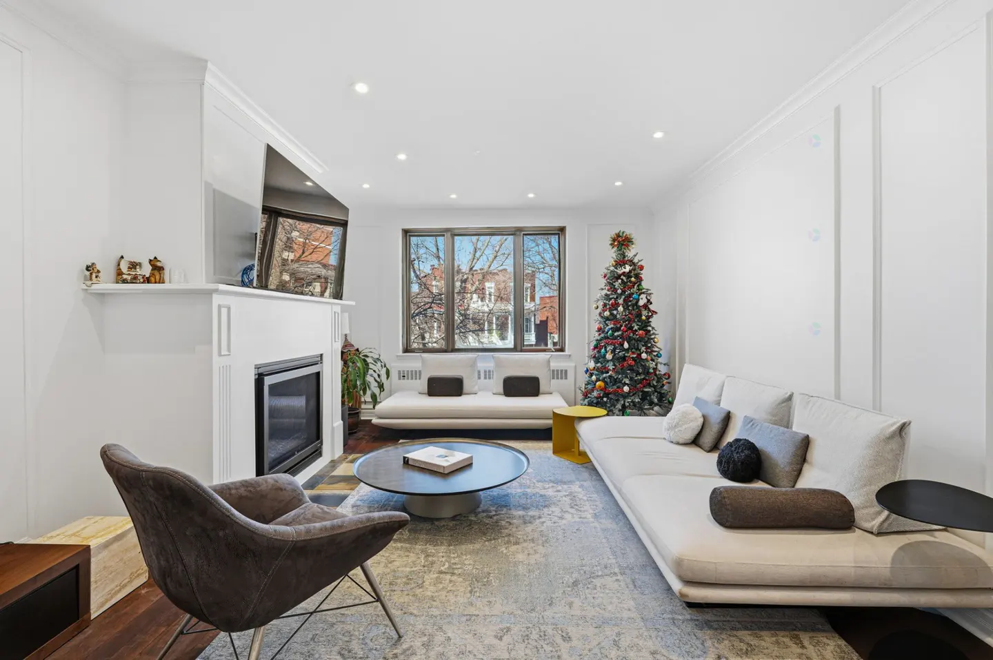 Living room with white walls, fireplace, TV, grey chair, and a Christmas tree near the window.