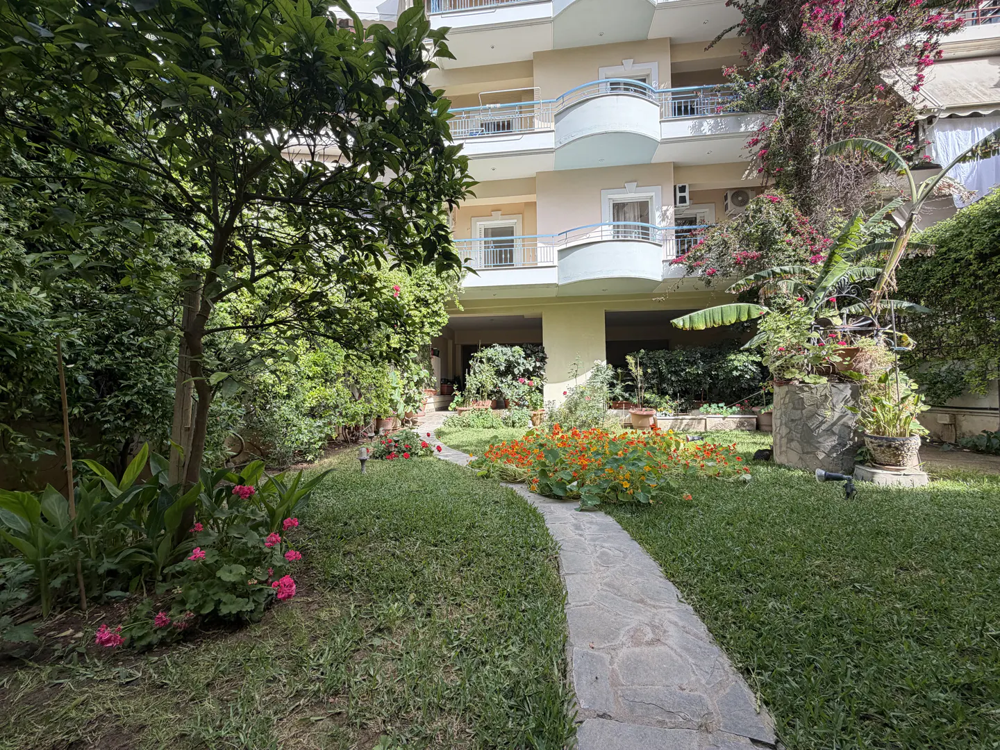 A stone path leads through a lush green lawn and colorful flowers to a light yellow apartment building with balconies.