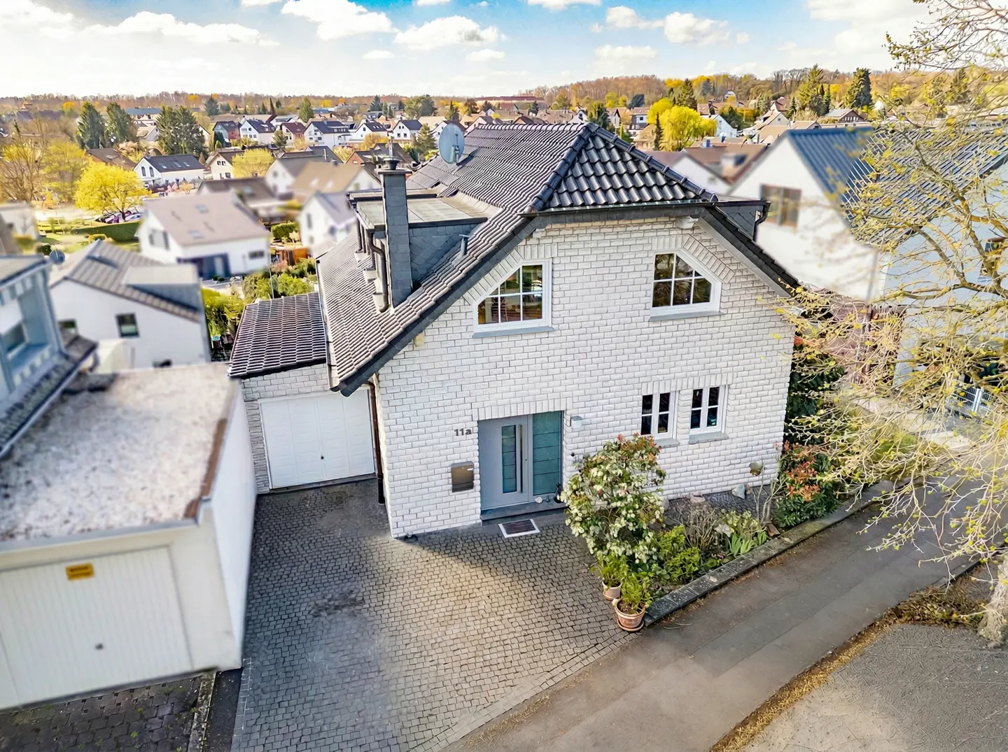 Aerial view of a two-story white brick house with a dark gray roof, a gray front door, and a cobblestone driveway.