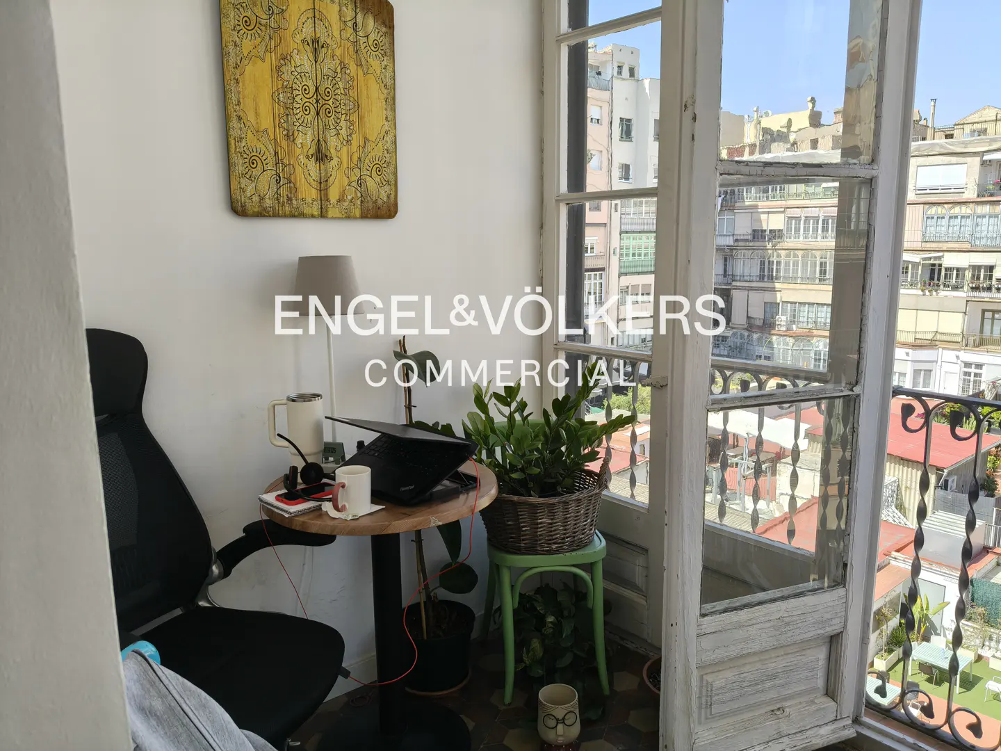 A small office space with a desk, chair, and open balcony with city views. A yellow patterned artwork hangs on the wall.