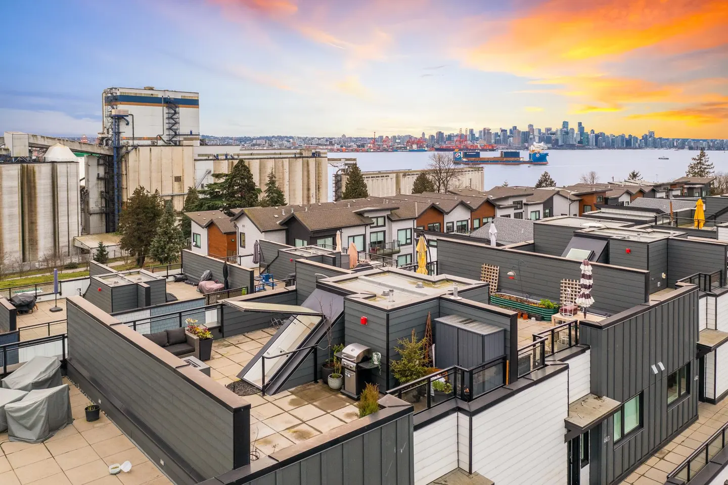 Aerial view of modern townhouses with rooftop patios, overlooking a cityscape and harbor at sunset.