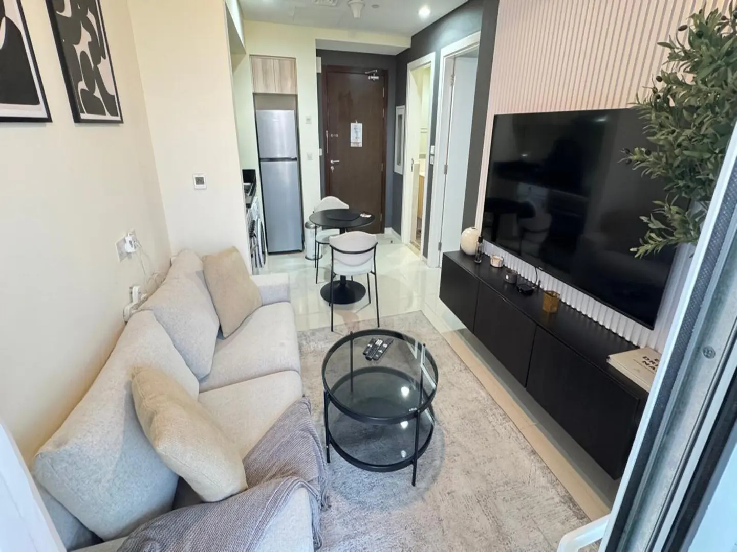 Living room with a beige sofa, glass coffee table, black TV stand, and a large TV. A dining table and chairs are visible in the background.